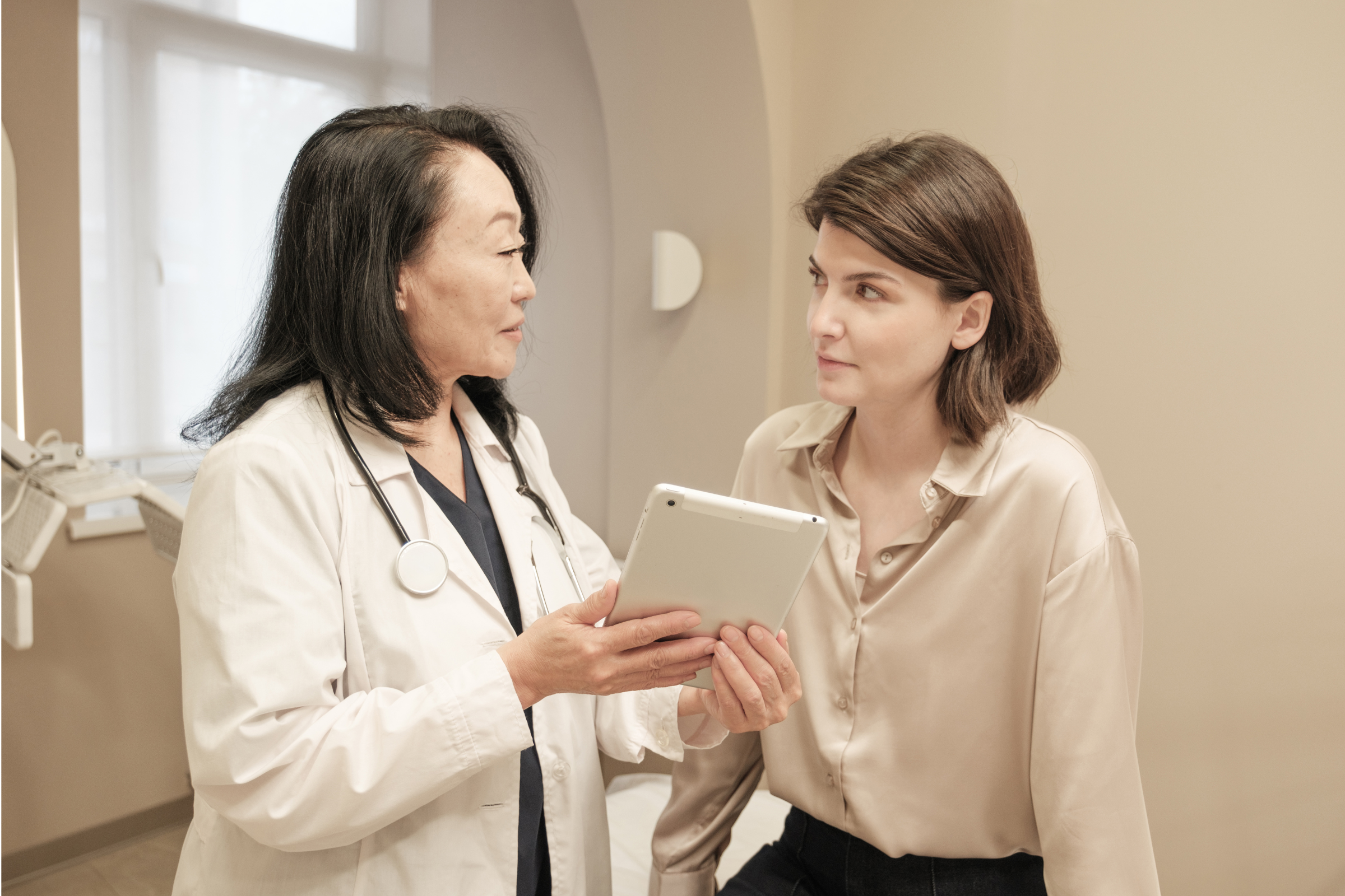 Woman with stethoscope around neck showing tablet device to another woman.